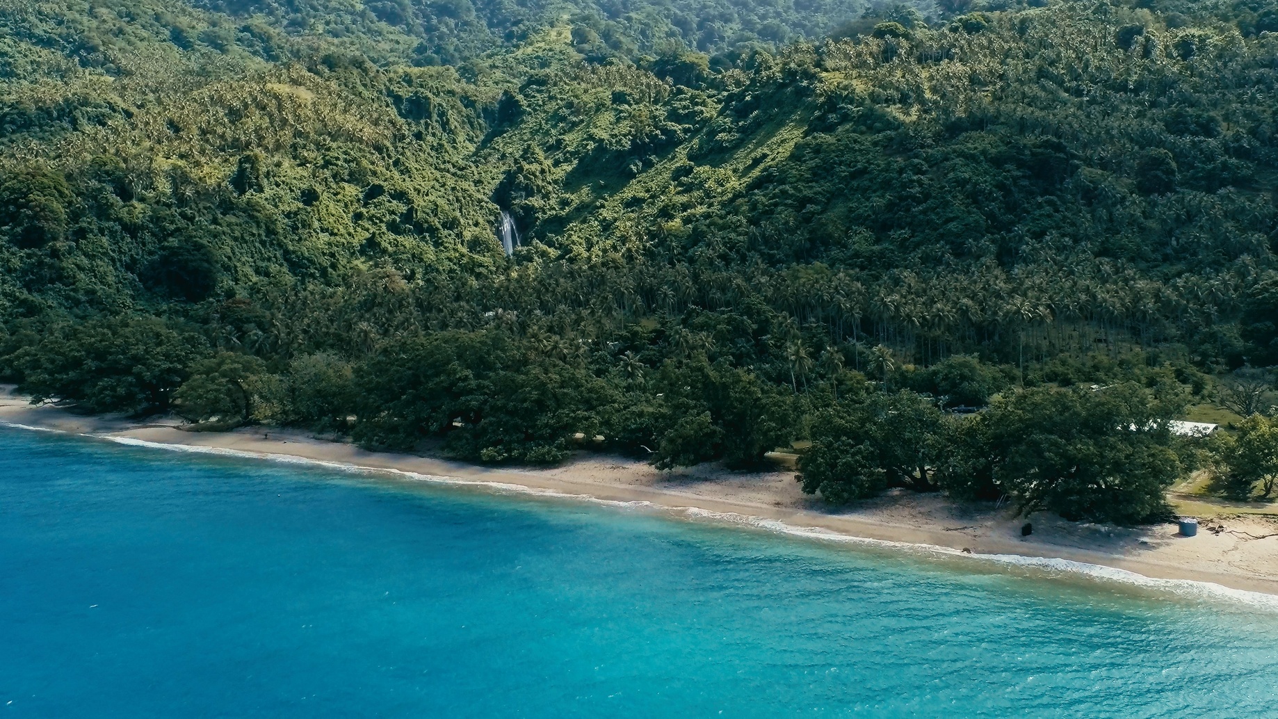 aerial drone image of a south pacific village on a remote island with sandy beach shore and lush tropical rainforest jungle with waterfall in the background